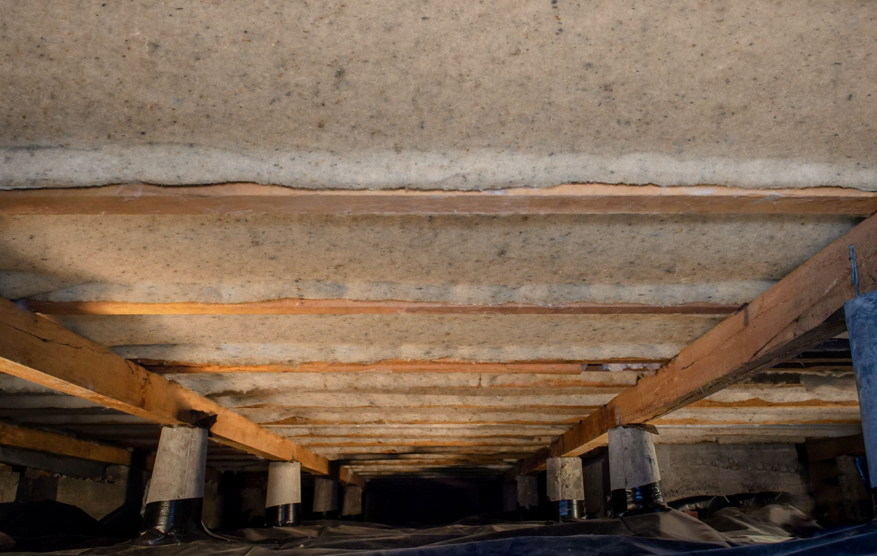 Retrofit underfloor insulation batts pinned between timber joists in a period-home subfloor, concrete stumps visible below